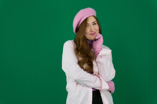 可爱的女孩戴着粉红色的法国帽子。年轻的女孩，头上戴着粉红色的贝雷帽，绿色背景上的法国时尚。不同的情绪粉色夹克手套棕色长发。粉红色的冬天图片下载
