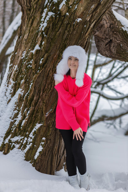 照片中，一位微笑的年轻女子身穿亮粉色毛衣，头戴带耳盖的白色蓬松帽子，在冬日的雪地公园里图片下载