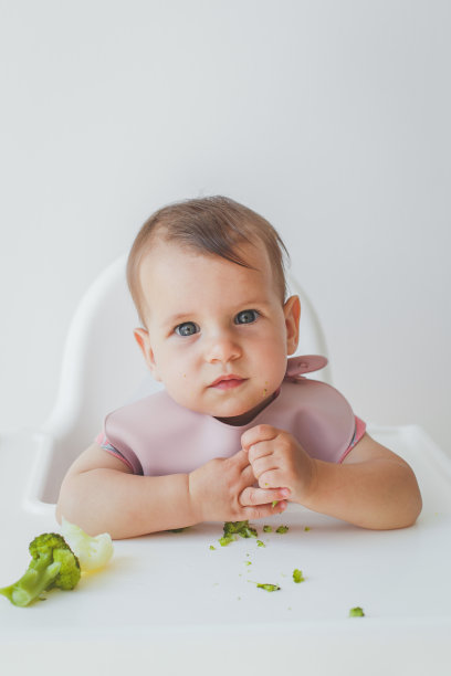 8个月大的女婴坐在高脚椅上，吃着辅食绿西兰花，特写镜头看着镜头。婴儿食品概念，垂直照片图片下载