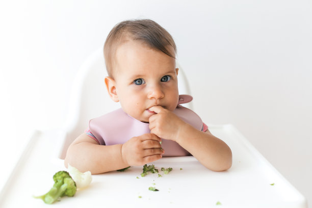 8个月大的女婴坐在高脚椅上，吃着辅食绿西兰花，特写镜头看着镜头。图片下载