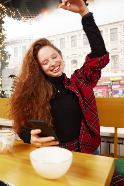 一个红发女孩拿着一杯咖啡，用智能手机拍照的照片。她看着手机的摄像头，微笑着图片下载