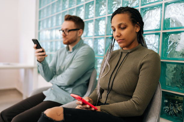 两个不同的年轻商人，招聘，使用手机在候诊室的网络，社交媒体或通信在办公室。图片下载