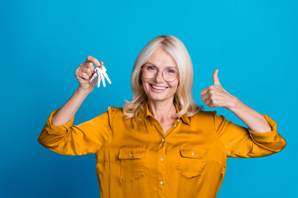 照片中，迷人的退休妇女手持一串钥匙，拇指朝上，穿着时尚的黄色绸缎衣服，衬托在蓝色的背景上图片下载
