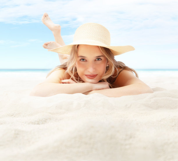微笑的女人躺在沙滩上看着镜头，戴着太阳草帽来保护她的皮肤。夏季沙滩度假和度假旅游的概念。海和蓝天在背景复制空间图片下载