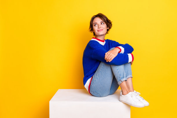 照片中甜美梦幻的女士穿着针织毛衣，坐在立方体上，回头看空旷的空间，孤立的黄色背景图片下载