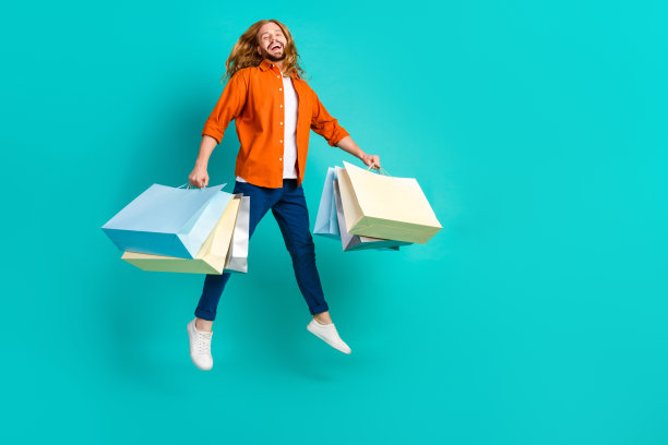 一个穿着橙色衬衫，手里拿着购物袋，在蓝绿色背景上跳跃的男人的全身照片图片下载