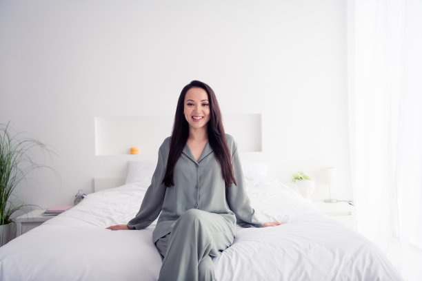 可爱的年轻女士的照片肖像坐在享受柔软的床单穿着时尚的灰色睡衣隔离在白色的卧室室内图片下载