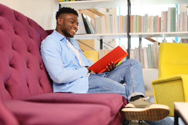 一个年轻人舒适地坐在现代化的图书馆里，沉浸在一本书中，在追求知识的过程中流露出自信和快乐。图片下载