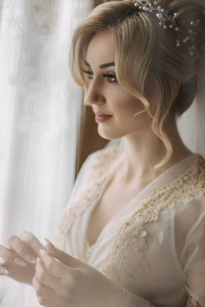 一位新娘梳着优雅的发型，头戴皇冠，身穿长袍，站在那里，拿着她的戒指看着它。美丽的妆容。婚礼的照片。金发女郎图片下载