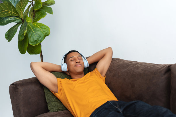 微笑的非洲裔美国年轻人戴着耳机，听着音乐，躺在舒适的沙发上图片下载