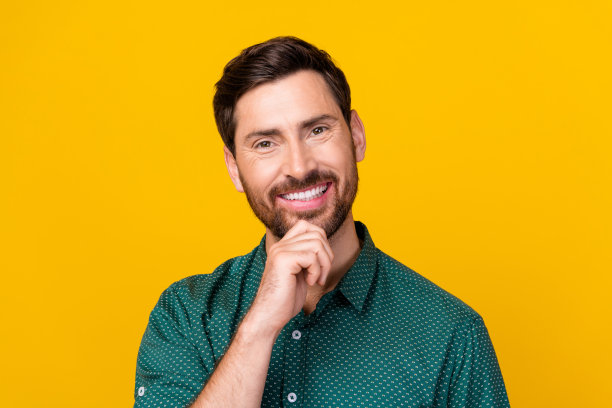 照片的愉快英俊的男人与深色头发穿着点衬衫手指在下巴上，牙齿微笑孤立的黄色背景图片下载