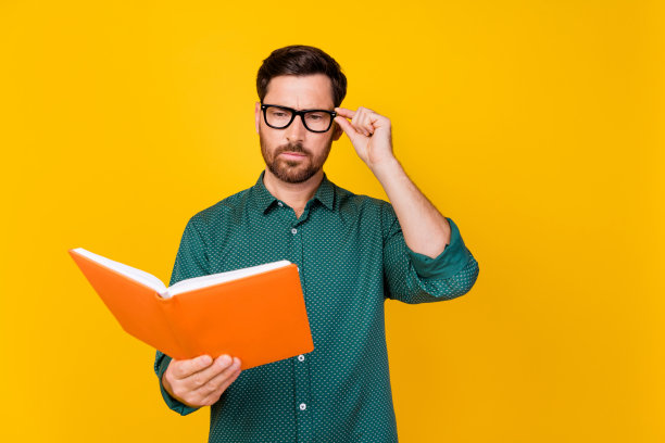 照片中的严肃聪明的男人，发型时髦，穿着带点的衬衫，戴着眼镜，在黄色的背景上读着书图片下载