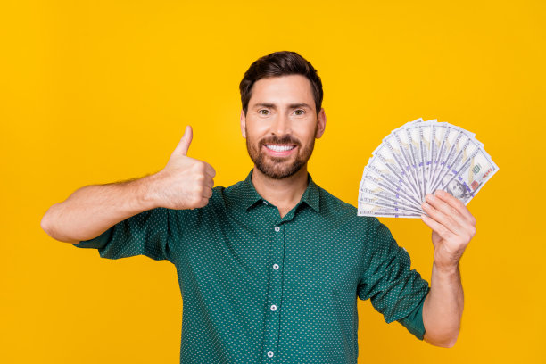 照片满意的积极的男人，时尚的头发，穿着点状衬衫，拿着美元，大拇指向上，孤立在黄色背景上图片下载