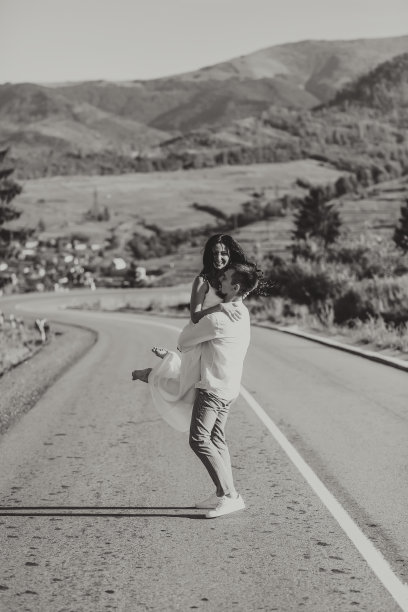 穿着白色衬衫的时髦新郎和身穿白色礼服的可爱黑发新娘在以森林和山脉为背景的柏油路上盘旋。黑白照片图片下载