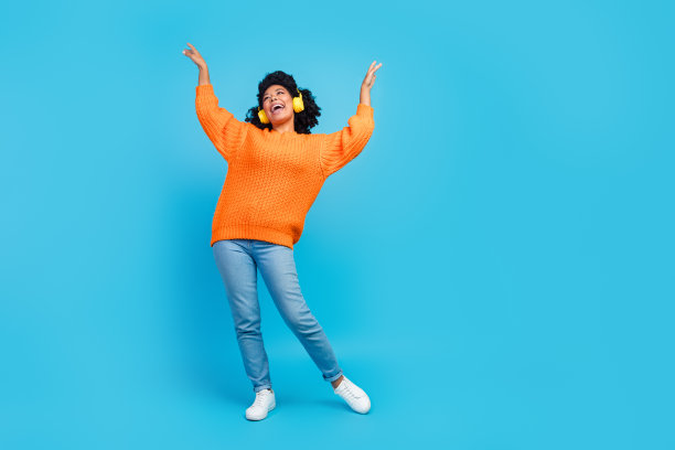 漂亮的年轻女孩跳舞的全尺寸照片，听收音机，穿时髦的针织衫，橙色的衣服，孤立的蓝色背景图片下载