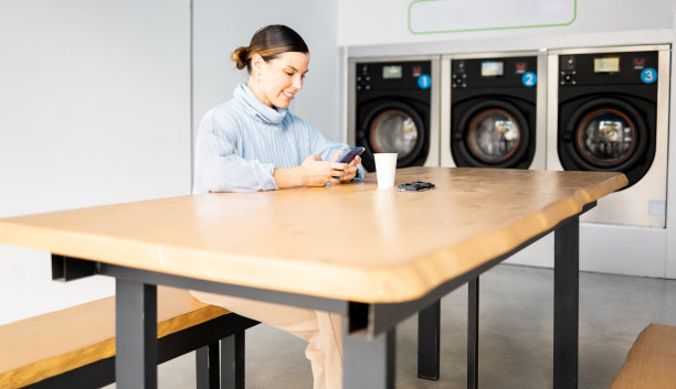 一个漂亮的女孩站在自助洗衣店里。这名女子坐在桌子旁等待洗衣服，同时她的注意力被手机分散了。公共自助洗衣机。图片下载