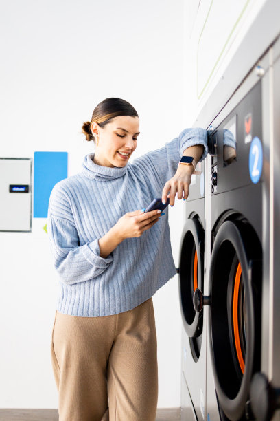 一个漂亮女孩站在自助洗衣店里的垂直照片。这名女子一边使用手机，一边靠在洗衣机上等待洗衣服。图片下载