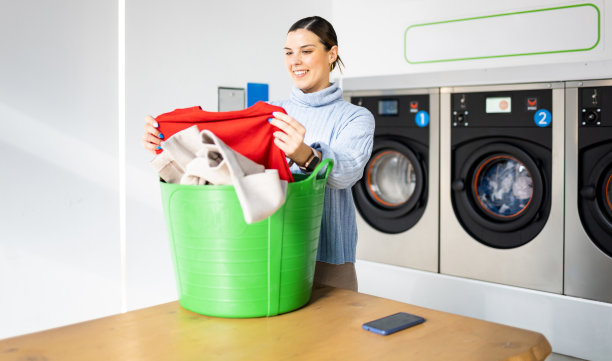 一个漂亮的女孩站在自助洗衣店里。一个女人在一个绿色的篮子里挑选衣服。公共自助洗衣机的概念。图片下载