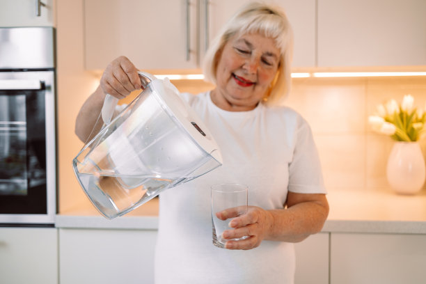 微笑的成熟的60岁妇女在厨房往玻璃杯里倒过滤过的水。补水，健康的生活方式。图片下载