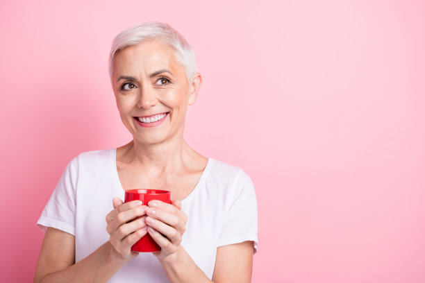 可爱的年长的女士的照片肖像拿着红色的咖啡杯，看起来空空荡荡，穿着时髦的白色衣服，孤立在粉红色的背景上图片下载