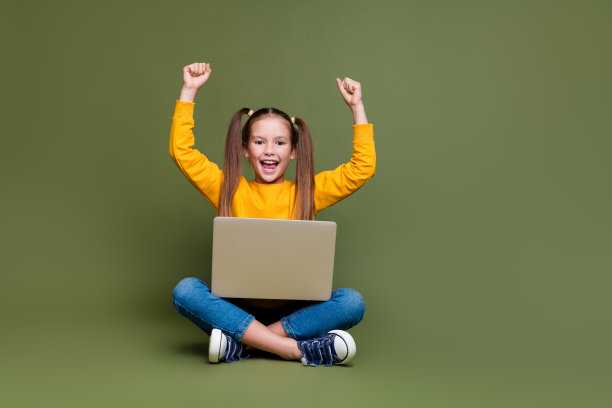 满意的小孩的全尺寸照片，穿着黄色套头衫，坐在笔记本电脑上，举起拳头，通过考试，孤立的卡其色背景图片下载