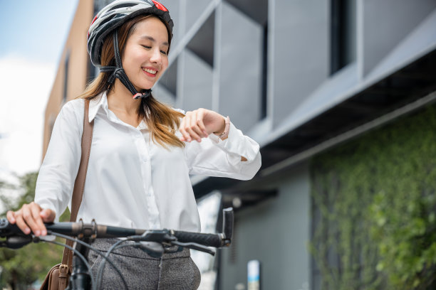 早上，一个年轻的女商人骑着自行车看时间，体现了商务通勤的生活方式。她职业的外表和头盔反映了她的职业。图片下载