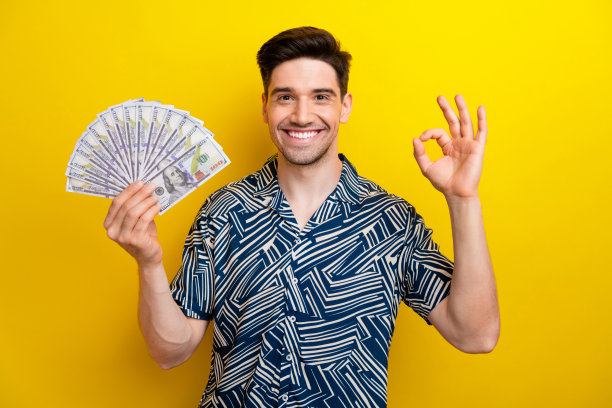 照片中富有的漂亮的年轻人，牙齿灿烂的微笑，手臂拿着美钞，在黄色的背景上展示了ok的标志图片下载