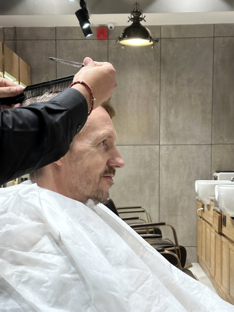 白人男子坐在美发沙龙的椅子上，穿着白色的理发师斗篷，无法辨认的人在剪头发，塑料黑色梳子，理发师的剪刀，侧面视图，重点放在前景图片下载
