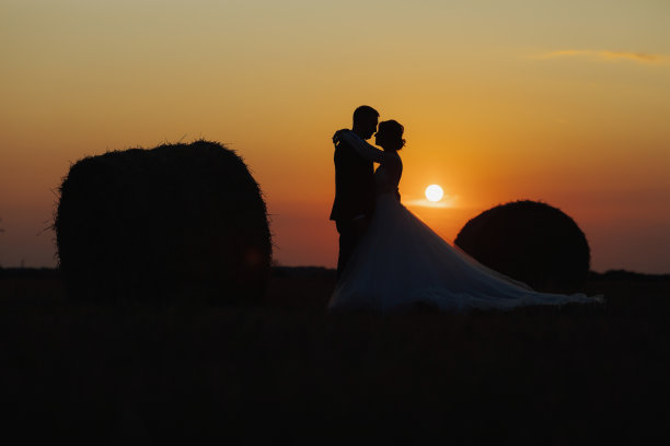 新娘和新郎的结婚照。日落时分，新郎新娘站在干草堆旁拥抱。身穿长裙的红发新娘。时髦的留胡子的新郎。剪影照片。图片下载