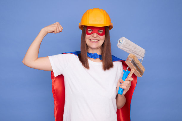 坚强快乐的女画家穿着超级英雄服装，戴着防护头盔，拿着绘画滚筒和画笔，孤立在蓝色背景上，举起手臂，展示她的二头肌和力量图片下载