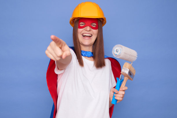 笑着的女画家穿着超级英雄的服装，戴着防护头盔，拿着绘画滚筒和画笔，孤立在蓝色背景上，指着相机选择你进行绘画工作图片下载