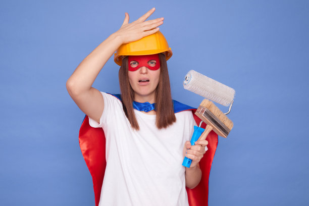 遗憾的是，不高兴的女画家穿着超级英雄服装，戴着防护头盔，拿着绘画滚筒和画笔，孤立在蓝色背景上，露出捂脸的手势，在油漆混合时犯了错误图片下载