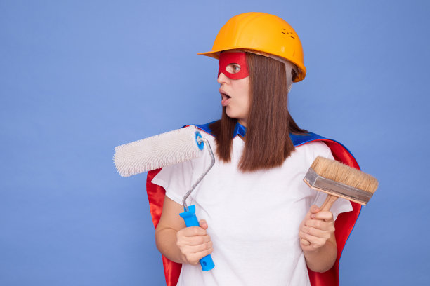 震惊的女画家穿着超级英雄的服装，戴着防护头盔，拿着绘画滚筒和画笔，孤立在蓝色的背景上，脸上带着惊讶的表情看着旁边图片下载