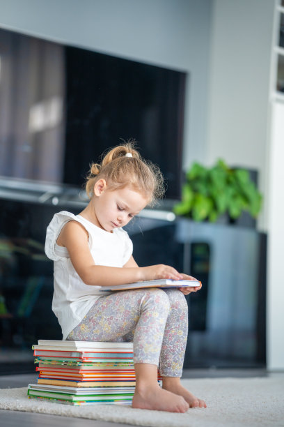 小女孩正坐在一叠儿童读物上，翻阅一本童话故事图片的书图片下载