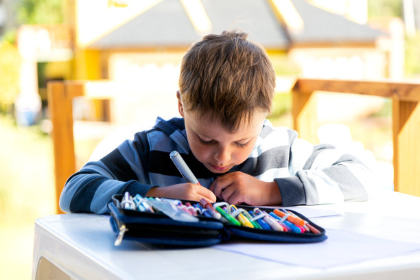 男孩用黑色马克笔画纸。他在户外坐在一张桌子旁。图片下载