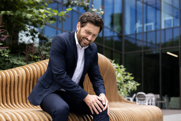 一位年轻的男性商人坐在办公中心附近街道上的长凳上，抱住自己的腿。愁眉苦脸，膝盖疼痛图片下载