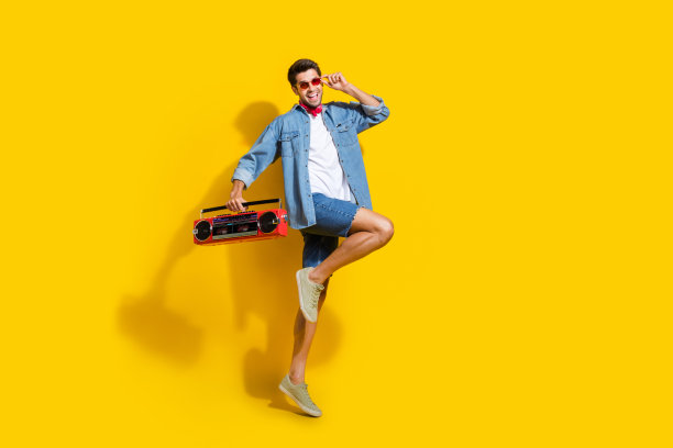 全身照片的酷男人穿着牛仔衬衫飞行触摸太阳眼镜持有音箱在聚会孤立的黄色背景图片下载