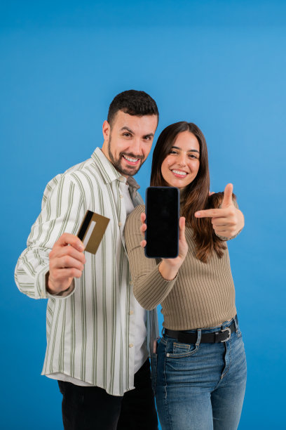 垂直的照片。一对年轻的成年夫妇，一男一女，在蓝色背景的工作室里摆姿势，他拿着信用卡，她指着智能手机。两个人在网上买相机图片下载