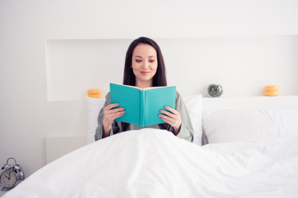 照片肖像迷人的年轻女子坐在床上看书睡前穿着时尚的灰色睡衣隔离在白色现代室内设计图片下载