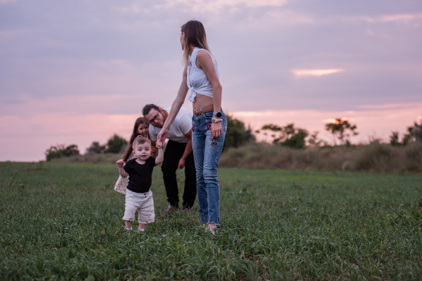 日落时分，一名妇女扶着蹒跚学步的孩子在草地上散步，背景是男人和孩子图片下载