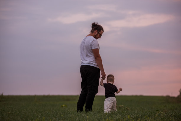 年轻男子在草地上牵着蹒跚学步的孩子的手，背景是柔和的夕阳天空图片下载