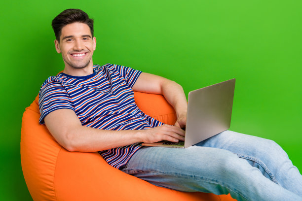 照片上的小伙子笑容满面，穿着条纹t恤牛仔裤，坐在沙发上，在绿色背景的笔记本电脑上写电子邮件图片下载
