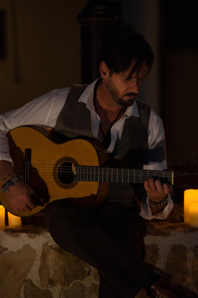 一个穿着黑白马甲的专业吉他手在夜晚的村庄广场上举行烛光音乐会的垂直照片。图片下载