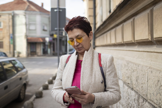 一位留着红色短发、戴着黄色太阳镜的多种族中年妇女，在镇上散步了很长时间。手里拿着智能手机。休闲优雅的风格。x世代。真实的人，下班后。春天开始的股票照片图片下载