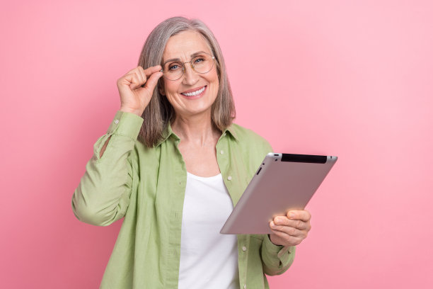 照片聪明的退休妇女触摸眼镜浏览集中现代平板电脑良好的生产力设备隔离在粉红色的背景图片下载
