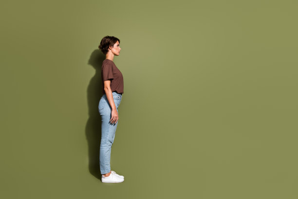 在卡其色的背景下，穿着棕色t恤牛仔裤的苗条愉快的女人的全尺寸侧面侧面照片看起来很空旷图片下载