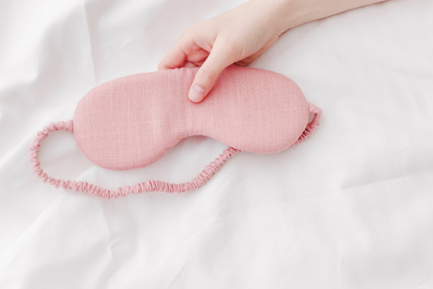 粉红色的眼罩睡在女人的手在白色的床上，最小的生活方式美学的照片。俯视图女性睡眠面膜，适合最佳睡眠者，适合旅行，舒适放松。好好休息的概念。图片下载