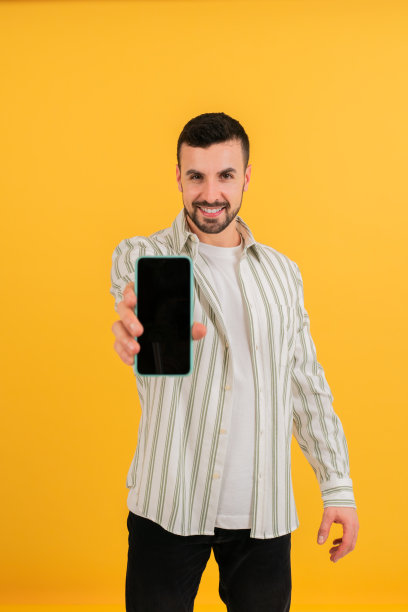 垂直的照片。一个年轻人微笑着在镜头前展示他的手机，微笑地站在一个孤立的黄色背景中。图片下载