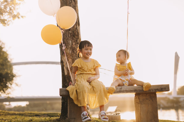 两个亚洲小女孩坐在花园公园的长椅上，手里拿着气球。库存图片图片下载