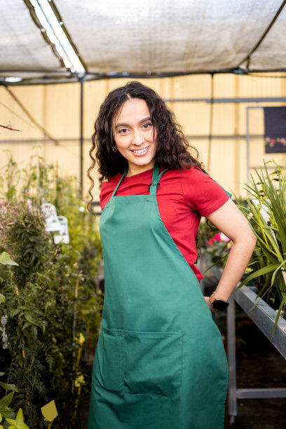 一个穿着深绿色围裙在托儿所工作的年轻拉丁妇女的垂直照片。这位非洲妇女一边系上围裙，一边开心地看着镜头。职业女性的概念。图片下载